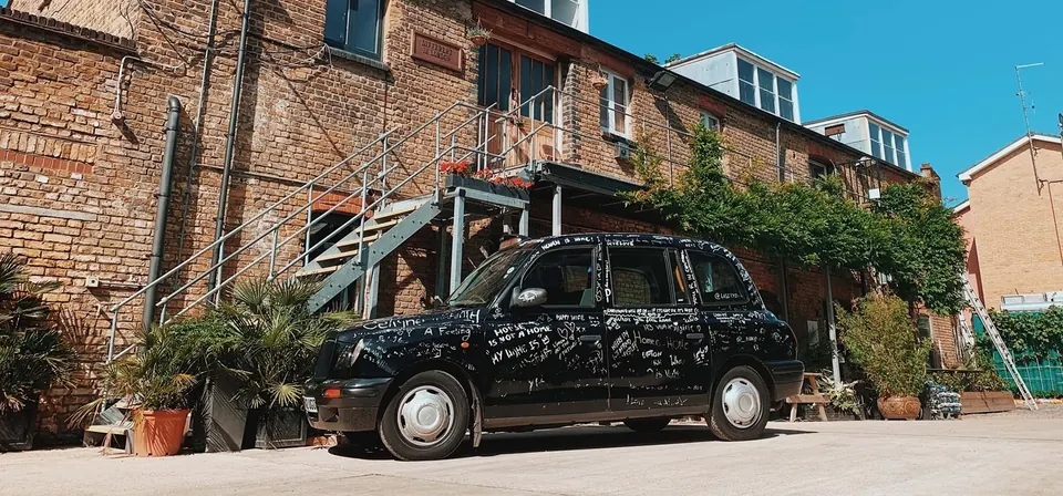 A black London cab covered in hand-written chalk messages, parked beneath the yard’s steel staircase