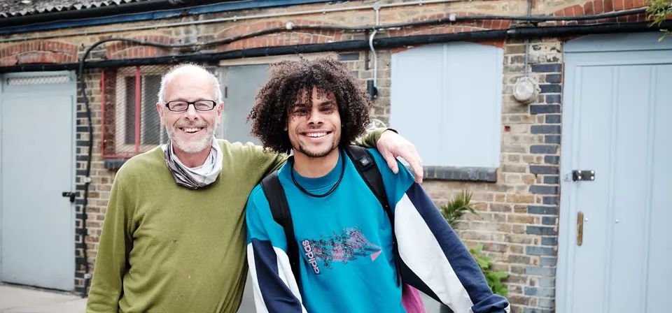 Two friends of the yard smiling, arm in arm, in front of the brick workshops