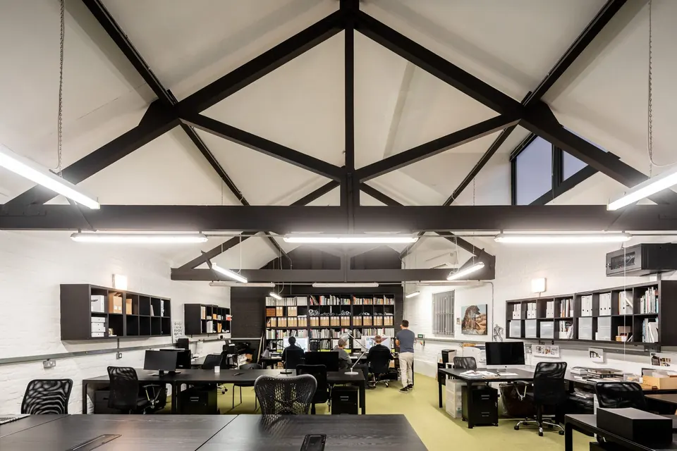 The coworking studio: a bright open-plan room under white roof trusses, with rows of desks and bookshelves